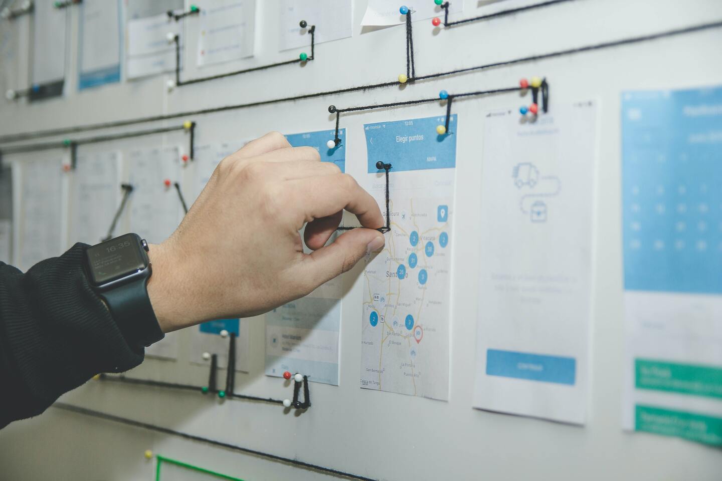 Eine Hand mit einer Smartwatch hält einen Pin an einer Karte auf einem Whiteboard mit verschiedenen Dokumenten und Diagrammen.