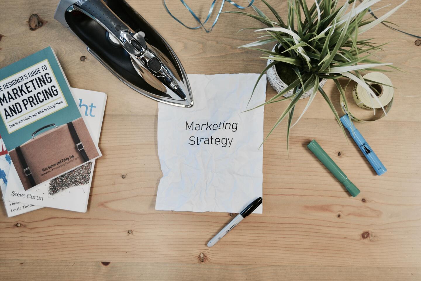 Ein Arbeitsplatz mit einem Notizzettel, auf dem "Marketing Strategy" steht, umgeben von Büchern, einem Bügeleisen, einer Pflanze und Schreibutensilien.