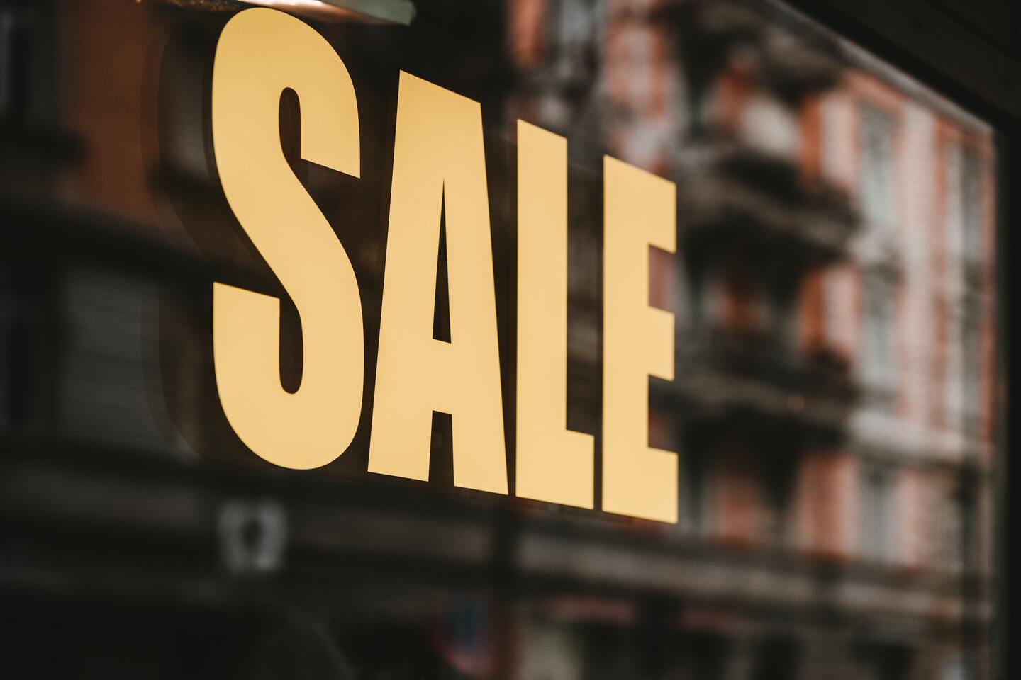 Ein großes gelbes "SALE"-Schild ist auf einem Fenster angebracht, mit unscharfem Hintergrund eines Gebäudes.